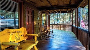 A wooden porch with a bench and rocking chairs, a wooden door, and a view of the surrounding forest.