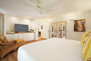 A bedroom with a large bed, a television on a cabinet, a wicker chair, and a ceiling fan.