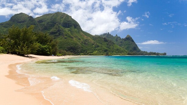 A beach with clear turquoise water, sandy shore, and lush green mountains in the background.