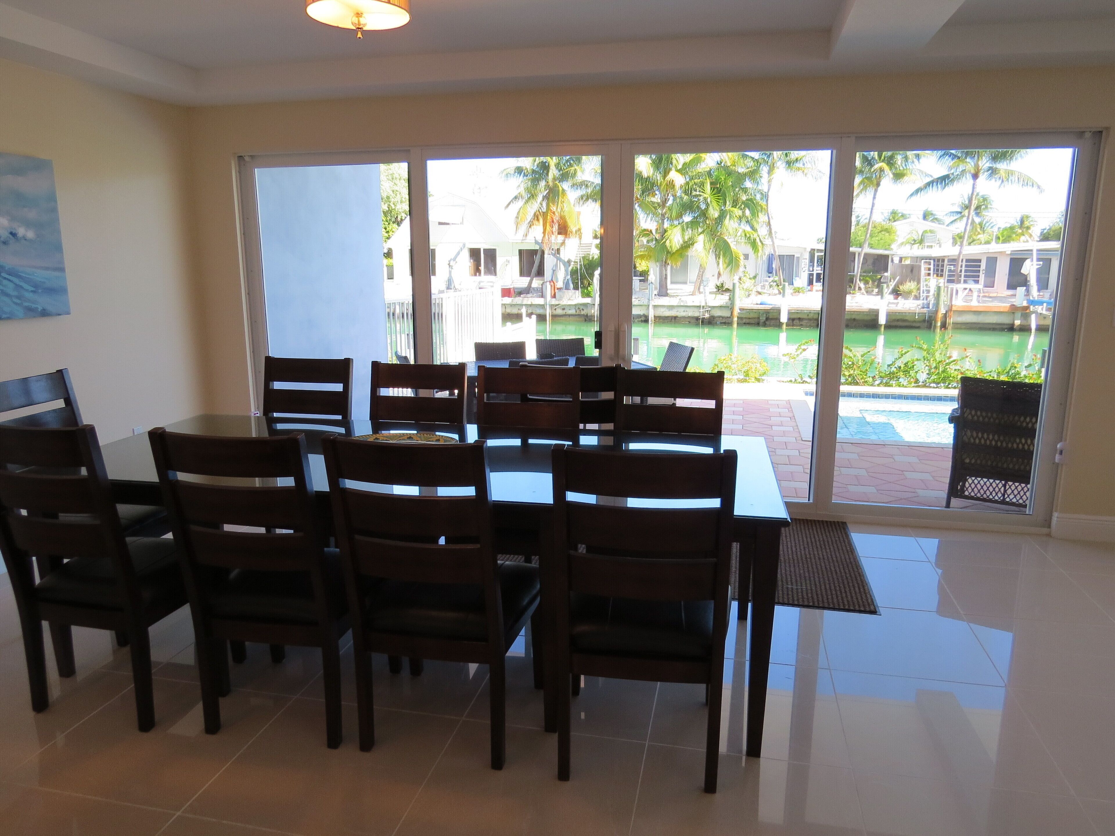 A dining area with a glass table and chairs, large windows with a view of a pool and palm trees, and a ceiling light fixture.