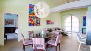 Un salón espacioso con una mesa de comedor de vidrio rodeada de sillas a rayas, una lámpara de araña blanca grande y obras de arte llamativas en las paredes.
