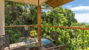 A glass-top table with wicker chairs on a balcony with a metal railing, surrounded by lush greenery.