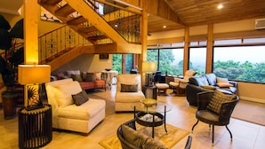 A spacious living area with a wooden staircase, comfortable seating, and large windows offering a view of the outdoors.