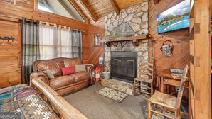 A cozy cabin interior with a stone fireplace, wooden walls, a leather sofa, and a television mounted on the wall.