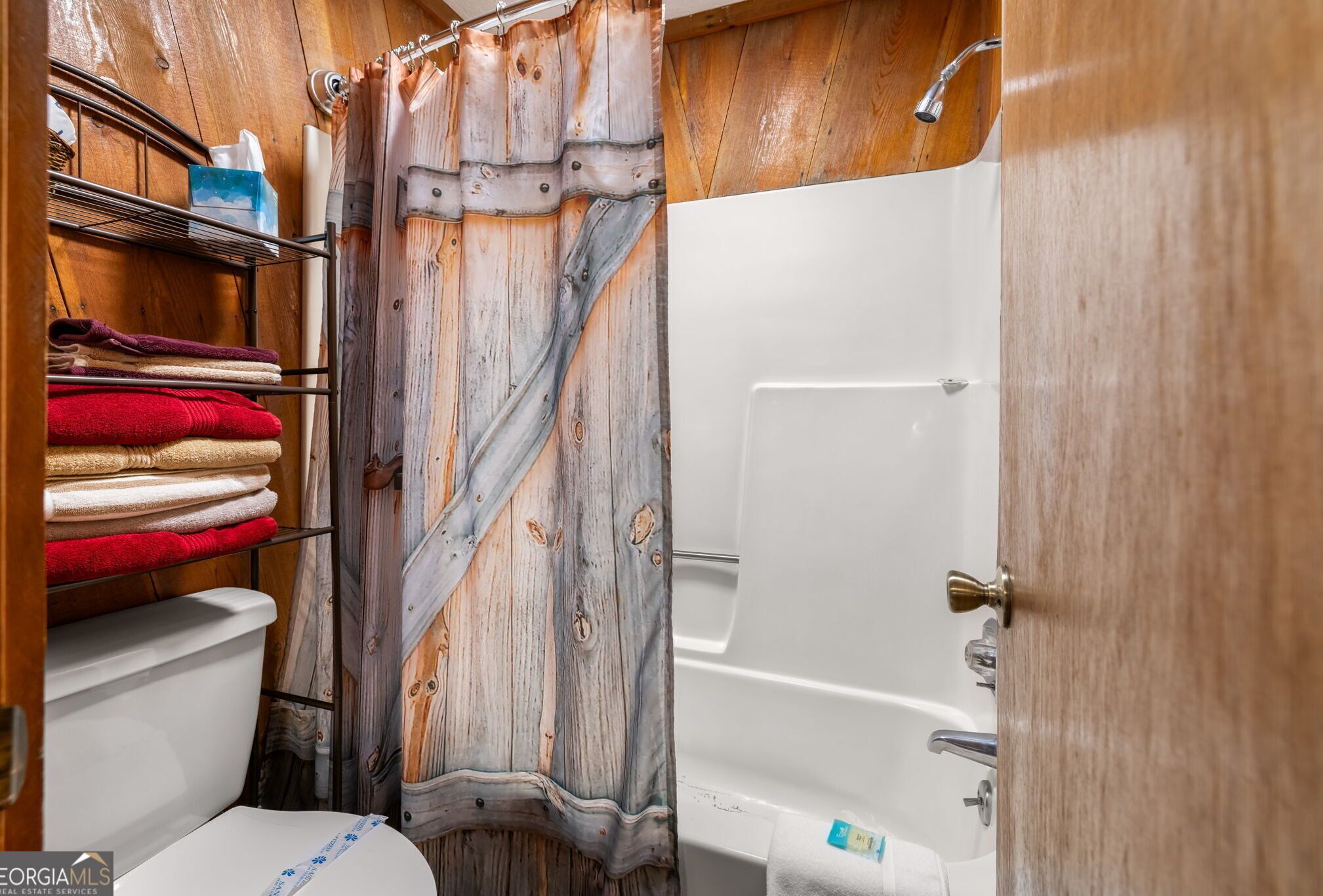 A bathroom with a rustic wooden theme, featuring a shower curtain, a toilet, and a shelf with folded towels.