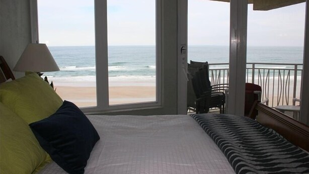 Un dormitorio con un ventanal que da a una playa, una cama con almohadas, una silla y una mesita.