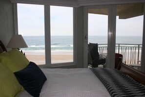 A bedroom with a large window overlooking a beach, a bed with pillows, a chair, and a small table. - Beautifully decorated, 3bed / 3BA - 5th Floor, Direct Ocean Front (Daytona Beach)