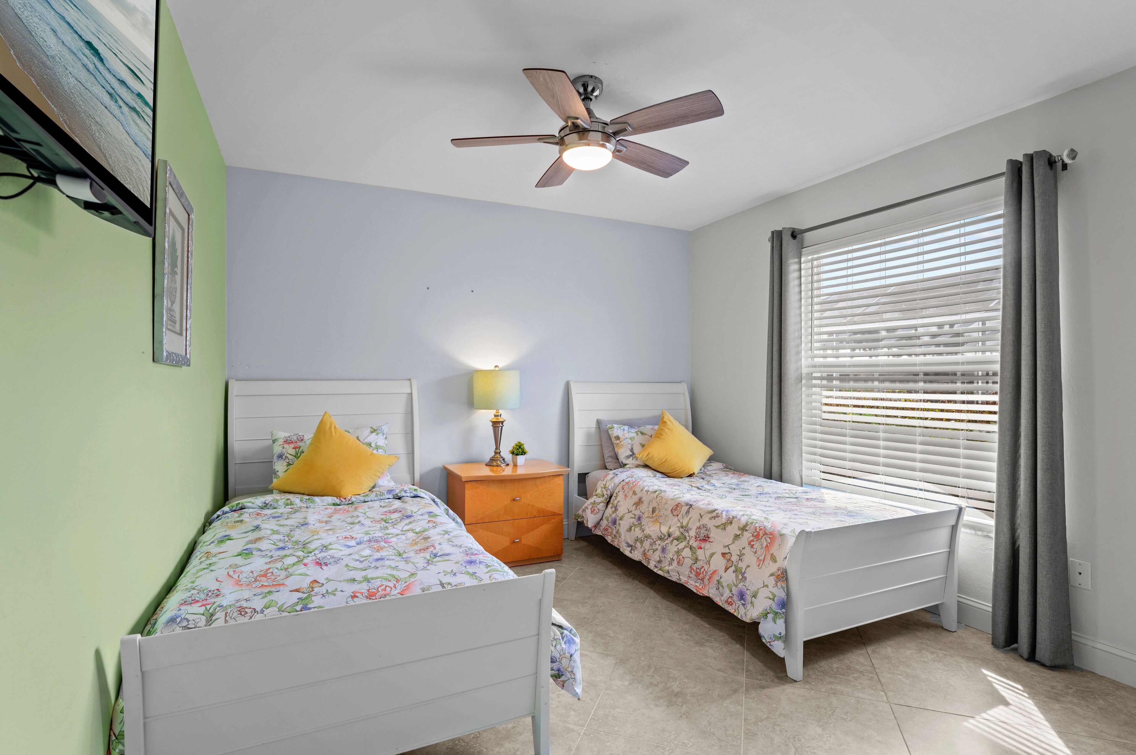 A bedroom with two single beds, a ceiling fan, a nightstand with a lamp, and a window with blinds.
