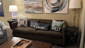 Un salon avec un canapé marron, deux tableaux accrochés au mur, une table d’appoint avec une lampe et une table basse avec des télécommandes.