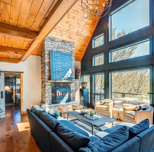 A spacious living area with a stone fireplace, large windows, and a high ceiling with wooden beams. - Owls Nest - Pool Table, hot tub, views, Indoor & Outdoor Fireplaces (Blowing Rock)