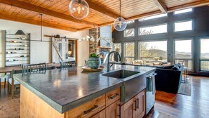 A spacious kitchen with a large island, wooden cabinets, and a stone backsplash. There is a dining area with a table and chairs, and a view of the outdoors through large windows. - Owls Nest - Pool Table, hot tub, views, Indoor & Outdoor Fireplaces (Blowing Rock)