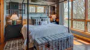 A bedroom with a four-poster bed, two bedside tables with lamps, a large window with a view of trees, and a woven basket on the floor. - Owls Nest - Pool Table, hot tub, views, Indoor & Outdoor Fireplaces (Blowing Rock)