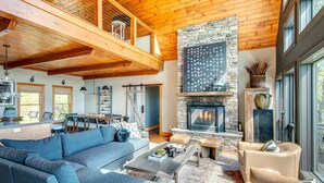 A spacious living room with a large blue sectional sofa, a fireplace, and a dining area with a wooden table and chairs. - Owls Nest - Pool Table, hot tub, views, Indoor & Outdoor Fireplaces (Blowing Rock)