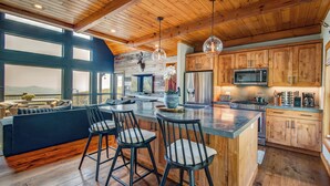 A spacious kitchen with wooden cabinets, a large island with bar stools, and a view of the mountains through large windows. - Owls Nest - Pool Table, hot tub, views, Indoor & Outdoor Fireplaces (Blowing Rock)