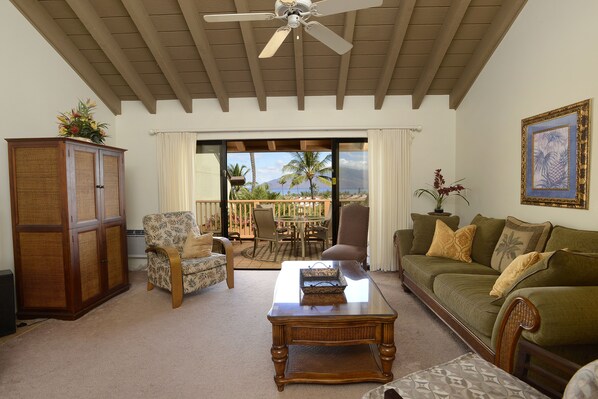 A living room with a ceiling fan, a wooden cabinet, a patterned armchair, a green sofa, and a view of palm trees through a sliding glass door.