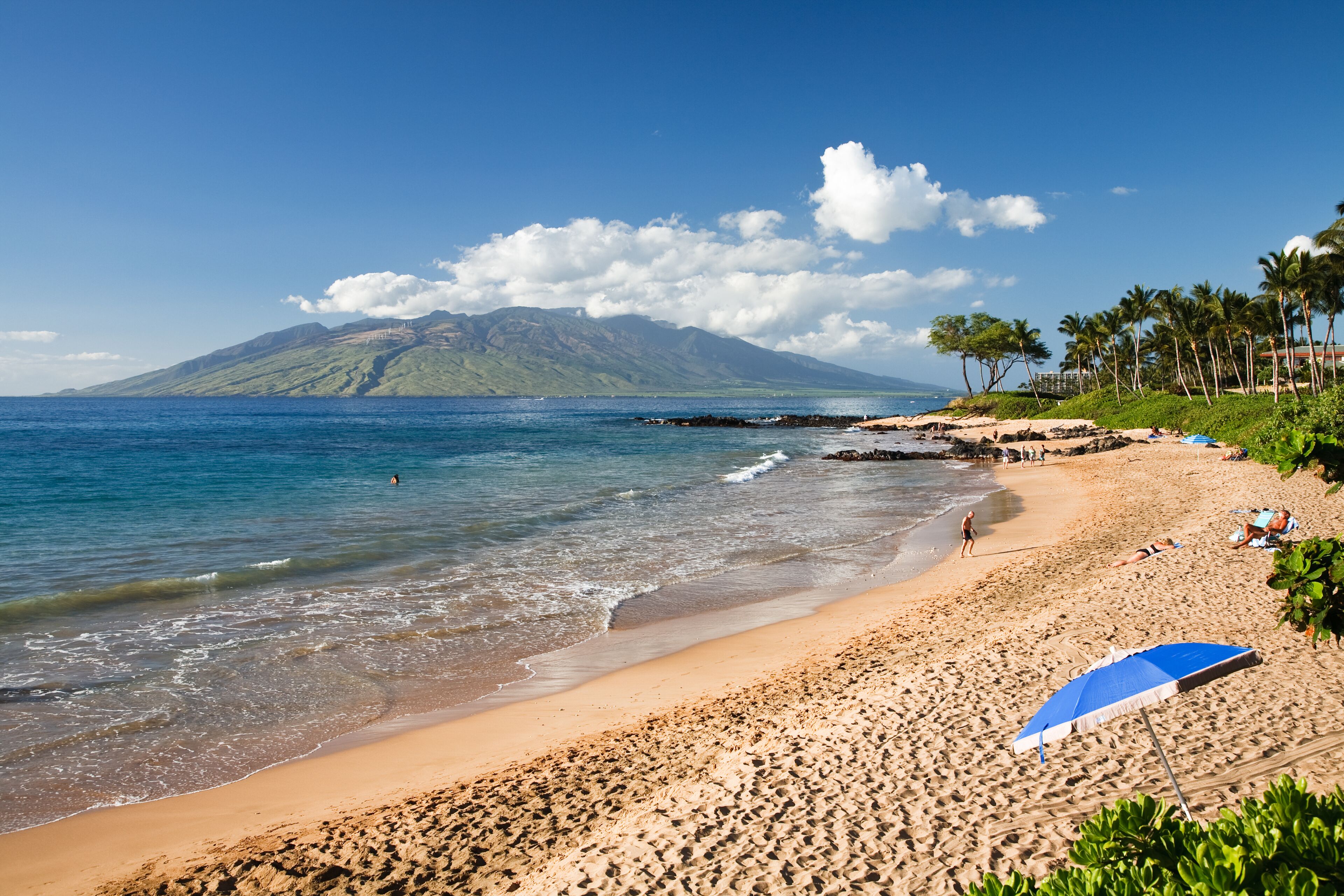 A beach with clear blue water, a sandy shore, and a mountain in the background.