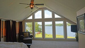 A bedroom with a large window overlooking a lake, a ceiling fan, and a television. - Luxury Lakefront Estate! 6BR 4.5BA, Private Dock,Ramp, Lakeside Firepit & More! (Greenwood)