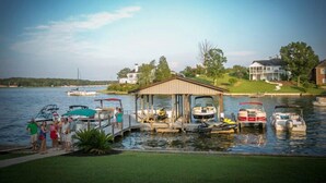 A lakeside dock with several boats, a covered structure, and a grassy area with houses in the background. - Luxury Lakefront Estate! 6BR 4.5BA, Private Dock,Ramp, Lakeside Firepit & More! (Greenwood)