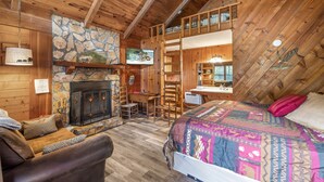A cozy cabin interior with a stone fireplace, wooden walls, a loft bedroom, and a desk area.