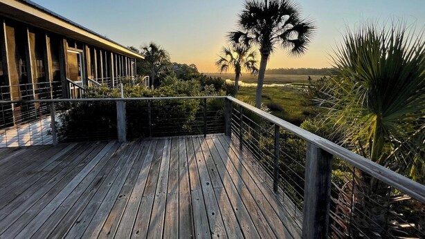 A wooden deck with a metal railing overlooking a landscape with palm trees and a building in the background. - Marshbreeze...on Tidal Creek with Marsh View (Darien)