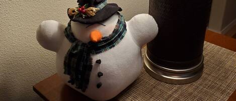 A snowman figurine on a wooden table next to a lamp.