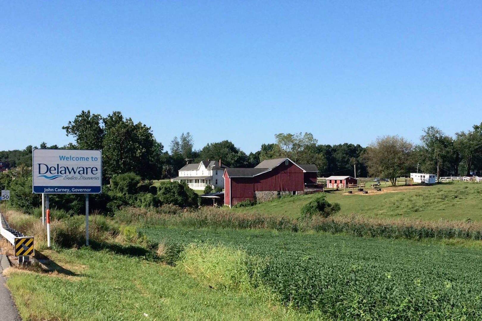 A rural landscape with a welcoming sign for Delaware, a red barn, and a white house.
