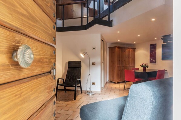 A modern interior with a wooden wardrobe, a black chair, a grey sofa, a dining table with red chairs, and a staircase.