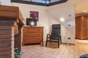 A room with a brick fireplace, a wooden dresser, a black chair, a floor lamp, and a colorful abstract painting on the wall.