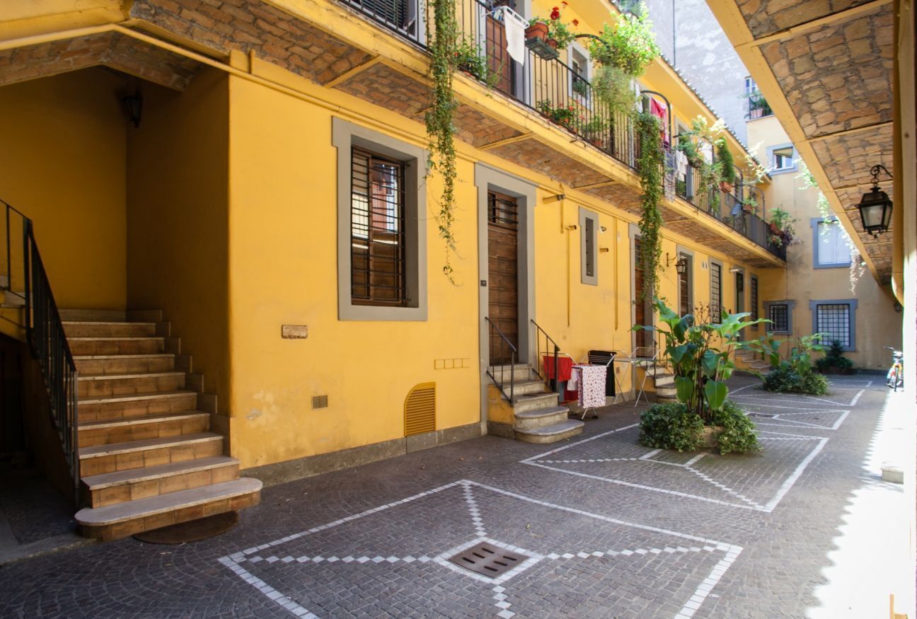 A yellow building with a staircase, a balcony with plants, and a cobblestone courtyard.
