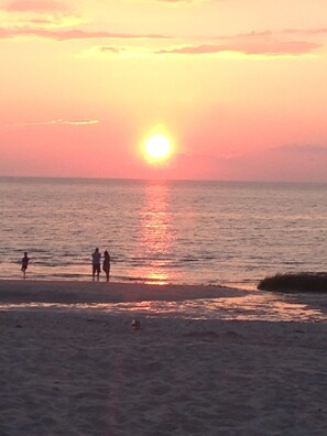 Un coucher de soleil sur une plage, avec des personnes debout près de l’eau.