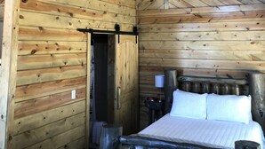 Interior de una cabaña de madera con una cama, una mesita y un reloj.