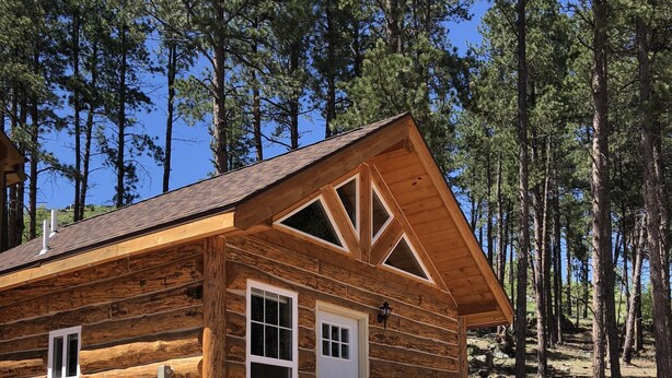 Una cabaña de troncos con terraza de madera y un portón de metal, rodeada de rocas y árboles.