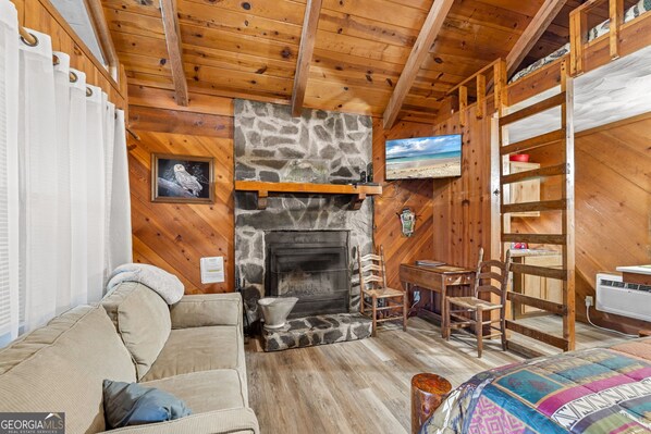 A cozy cabin interior with a stone fireplace, wooden walls, a sofa, and a bed. - Bear Paw Cottage #5  Queen Log Bed: COZY!: Pet Friendly; 2 Adults/2 Children (Sautee Nacoochee)