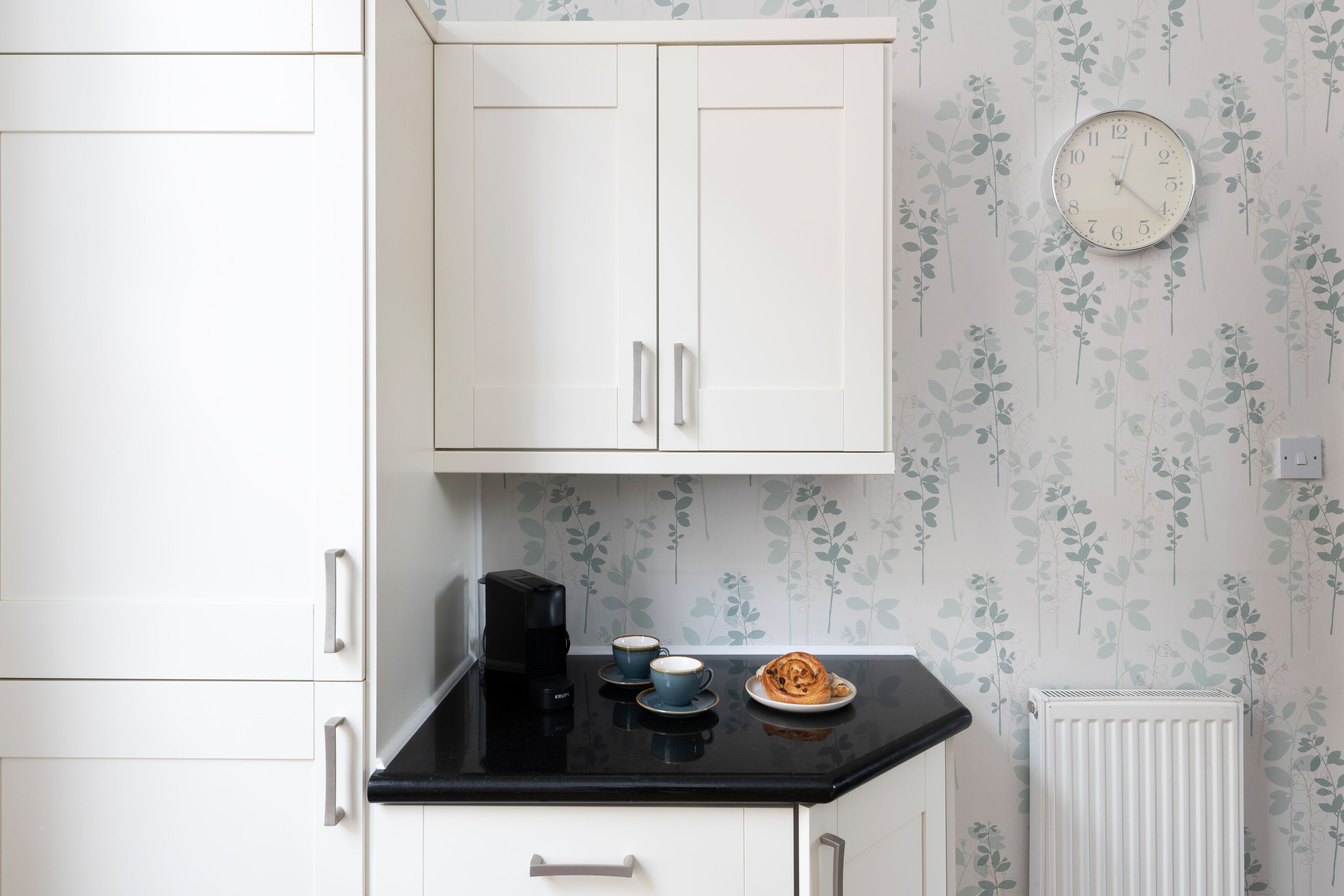 A kitchen with white cabinets, a black countertop, a coffee maker, two cups, and a plate with food.