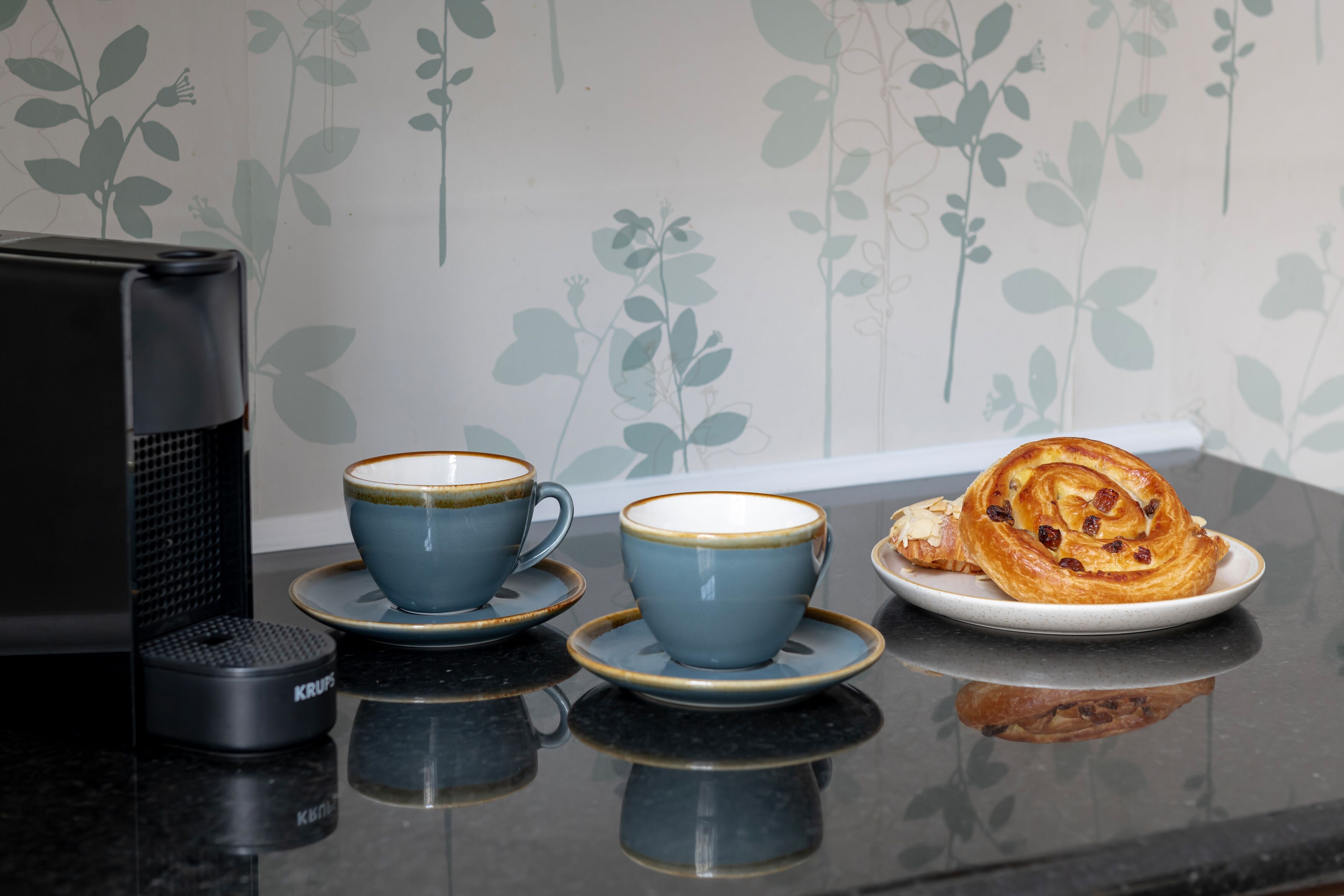 Eine Küchenarbeitsplatte mit zwei blauen Tassen, einem Teller mit einem Gebäck und einer Kaffeemaschine.