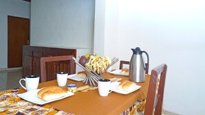 A dining table with a brown tablecloth, white plates, a coffee pot, and a banana stand. - Feel At Home (Kigali)