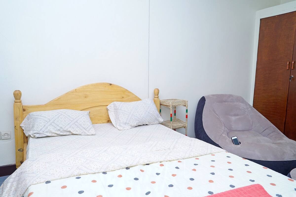 A bed with a wooden headboard, two pillows, a polka dot bedspread, a wicker side table, and a gray chair.