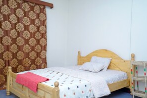 Un lit simple avec une tête de lit en bois, une couvre-lit blanche à pois et une couverture rouge. Une table de chevet en bois avec un panier tressé se trouve à côté du lit.