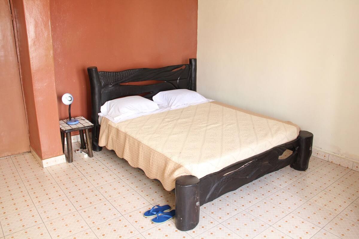 A single bed with a beige bedspread and white pillows, a small bedside table with a lamp, and a pair of blue slippers on the floor.