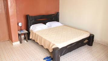 A single bed with a beige bedspread and white pillows, a small bedside table with a lamp, and a pair of blue slippers on the floor.