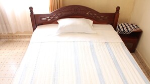 A neatly made bed with a striped bedspread, a wooden headboard with floral carvings, and a bedside table with a patterned cloth. - Joy Guesthouse (Kigali)