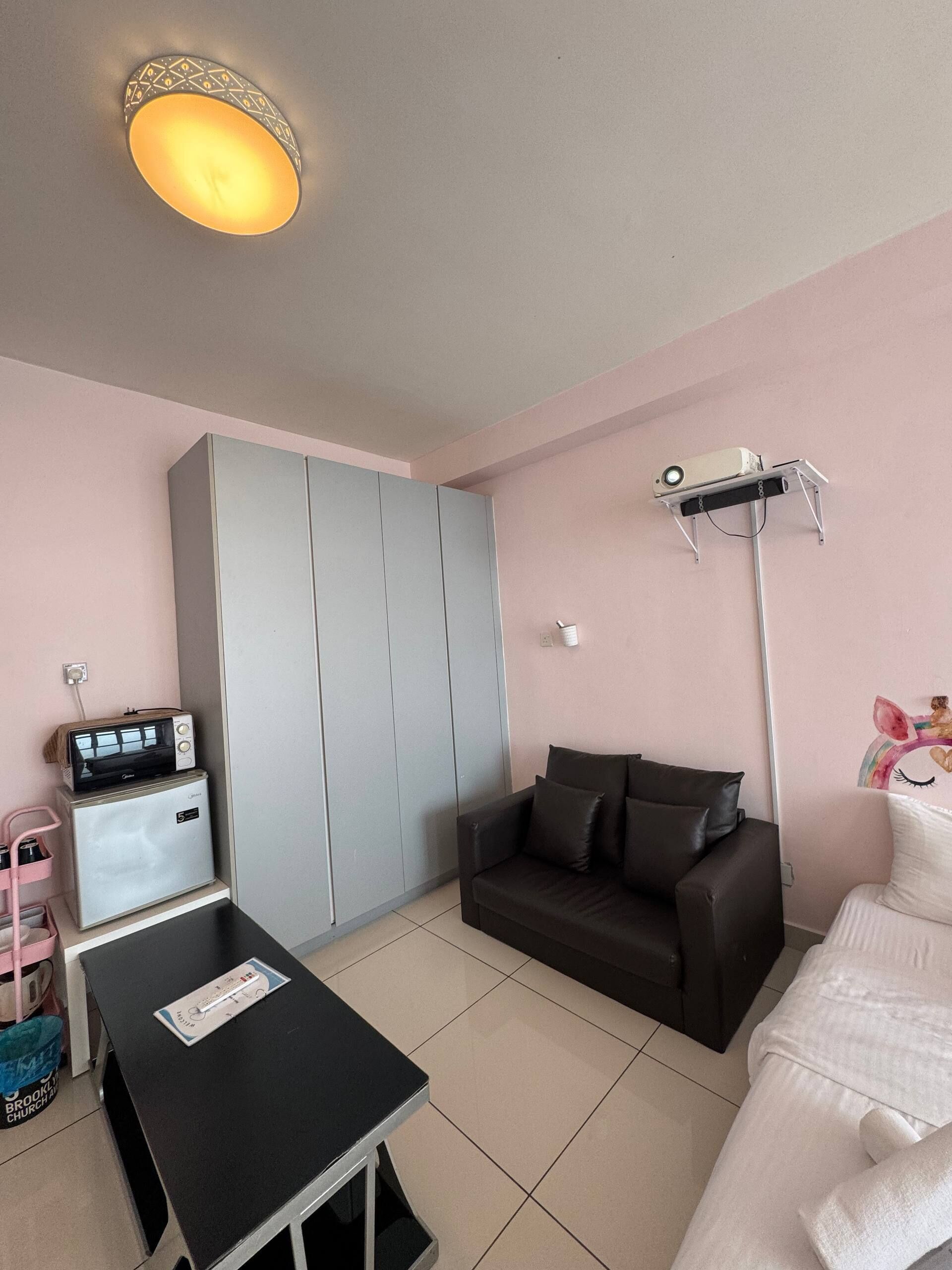 A room with a kitchenette, a sofa, a coffee table, a ceiling light, and a projector mounted on the wall.