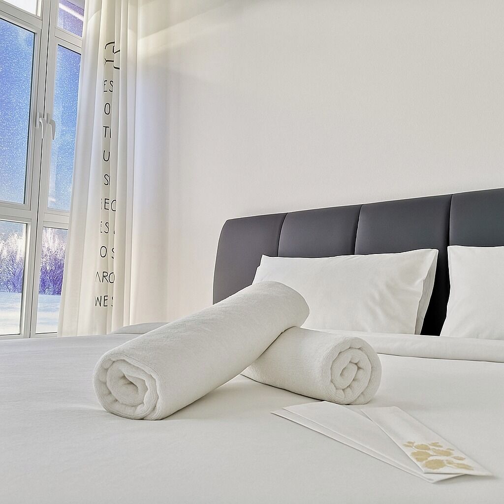 Una cama bien hecha con sábanas blancas y toallas enrolladas, un cabecero oscuro y vistas a árboles a través de una ventana.