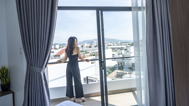 A woman standing by a balcony with a city view, curtains on either side, and a potted plant on a table. - 7S Hotel Hoang Anh & Apartment (Nam Nha Trang)