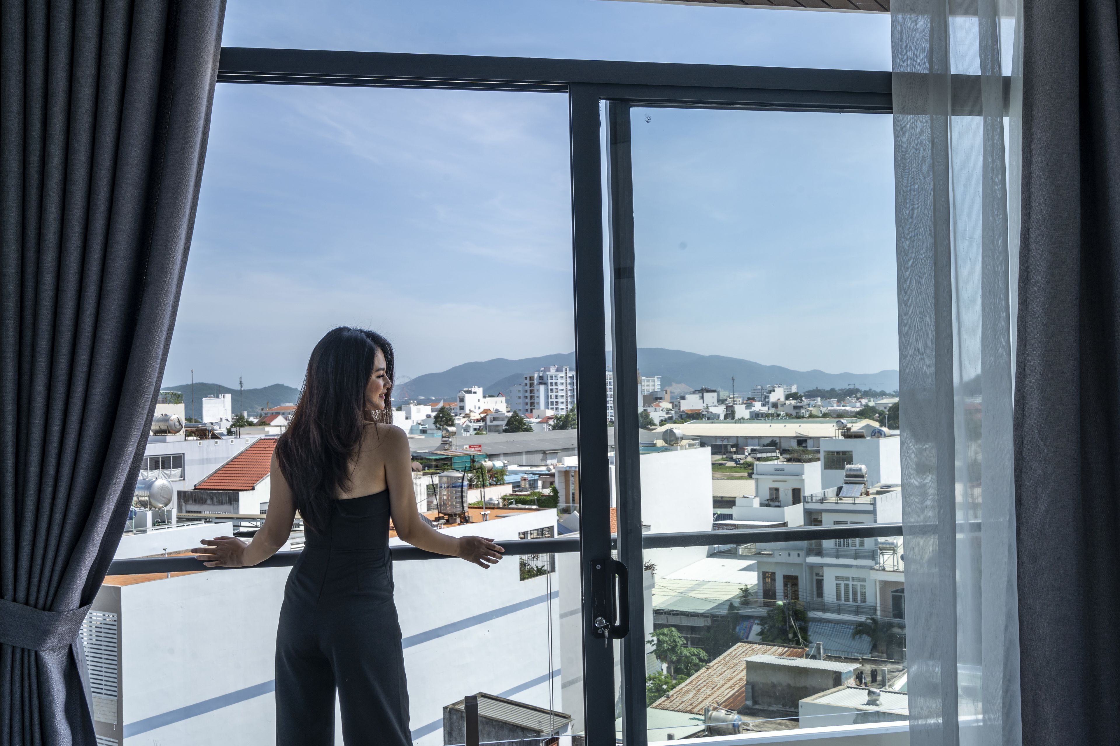 Une personne se tient près d’une grande fenêtre, d’où l’on peut admirer un panorama sur la ville, avec des bâtiments et des montagnes visibles au loin.