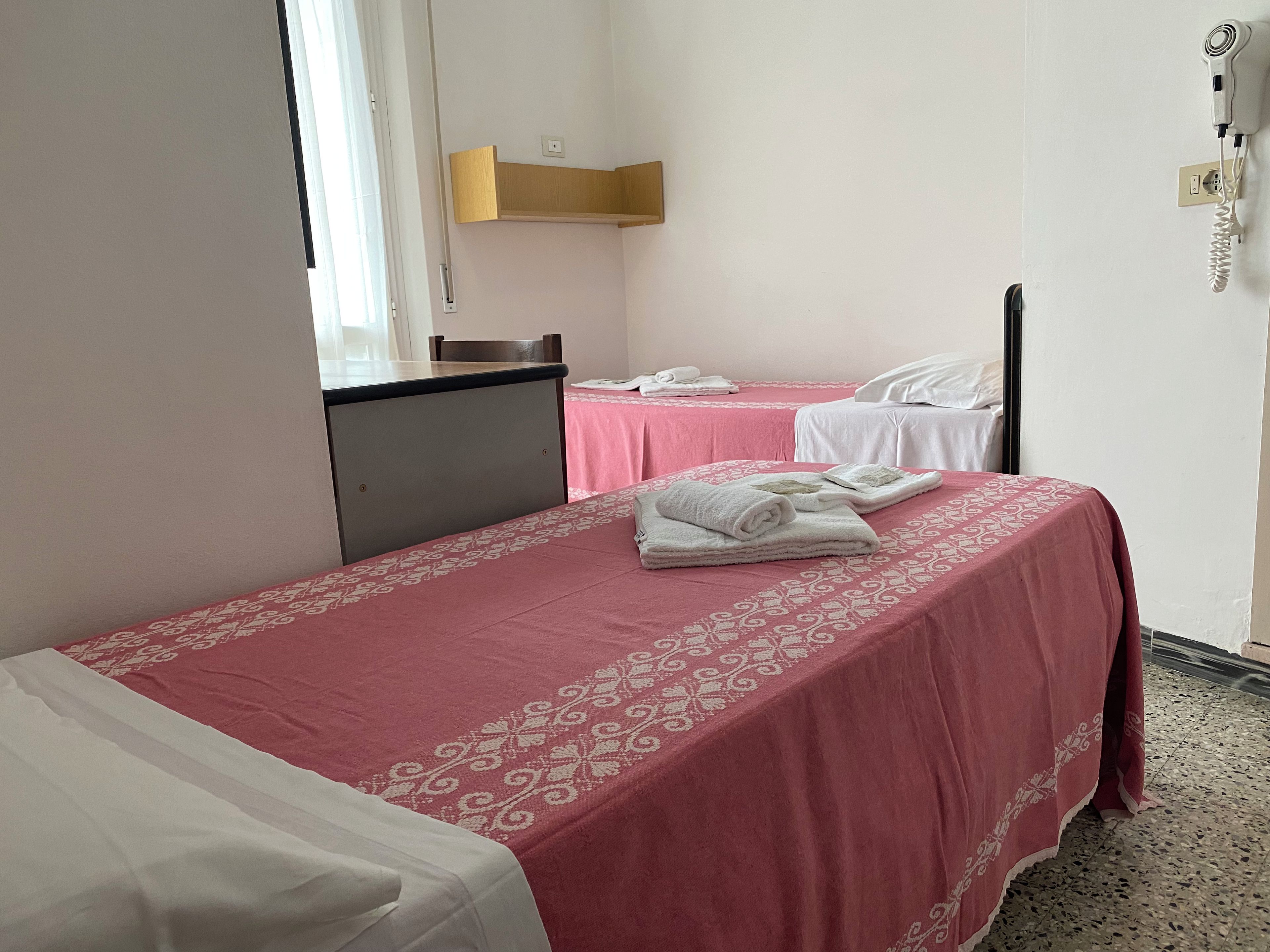 A hotel room with two beds, each with a pink bedspread and white linens. There is a desk with a chair, a small table with a pink tablecloth, and a wall-mounted phone.