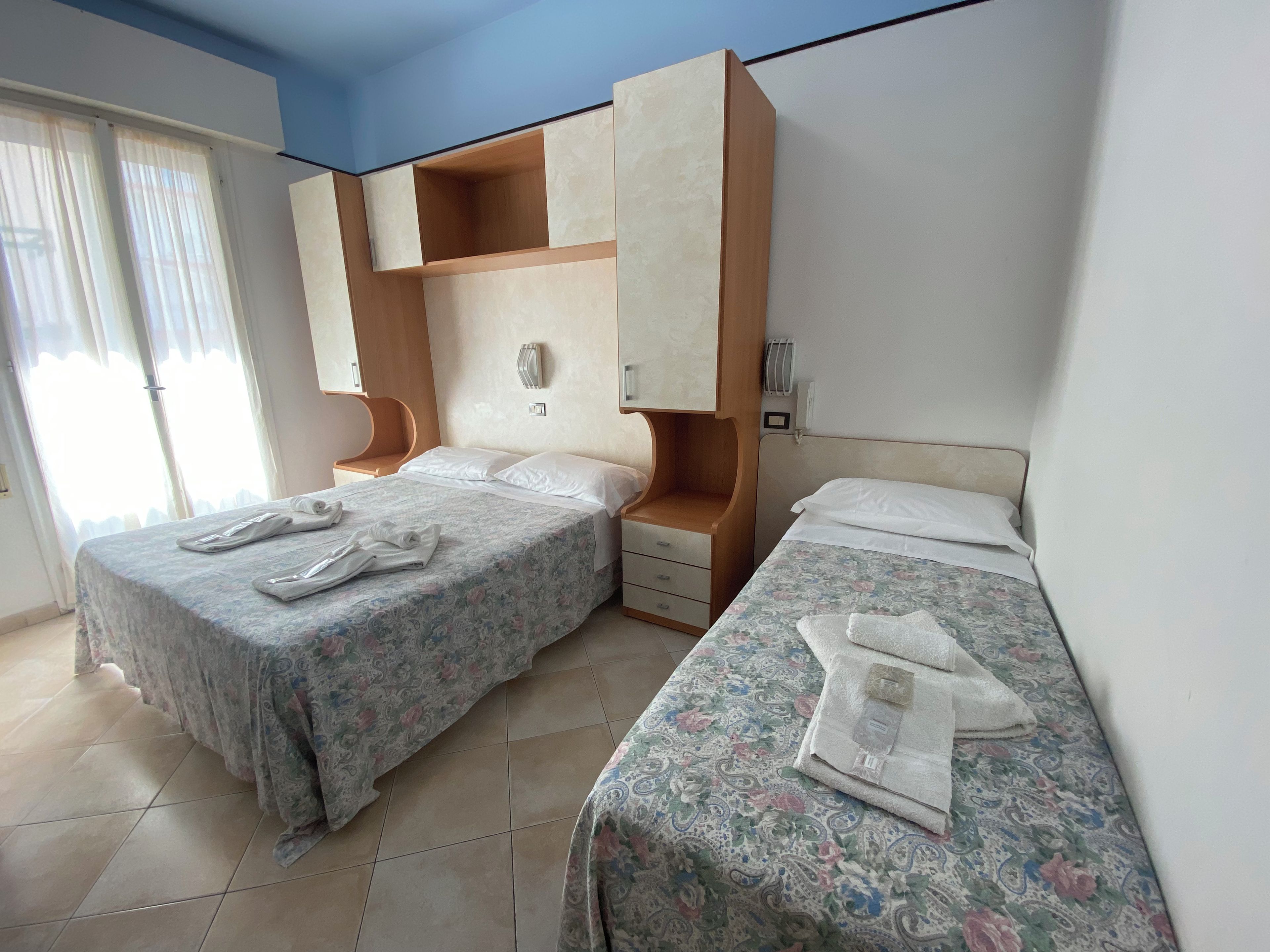 A hotel room with two beds, a wooden wardrobe, a bedside table, and a window with curtains.