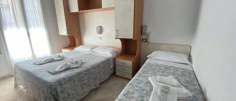A hotel room with two beds, a wooden wardrobe, a bedside table, and a window with curtains.
