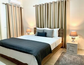 A hotel room with a bed, two bedside lamps, a wooden headboard, curtains, and a small nightstand with drawers. - Elite Residence (Btouratij)