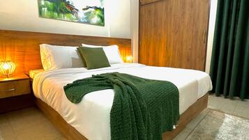 A neatly made bed with white linens and a green throw blanket, a wooden headboard, a nightstand with a lamp, and a painting on the wall.